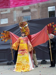 Eve wearing a gown under a large paper mache mask