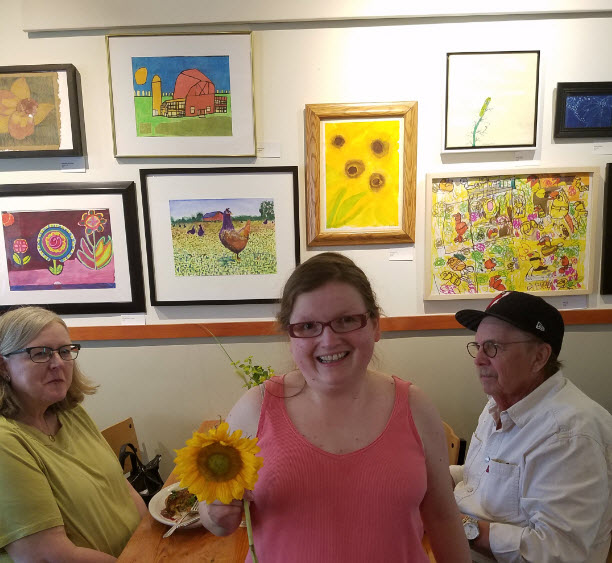 Alt Text = Evita in orange top, holding a sunflower, standing by sunflower painting